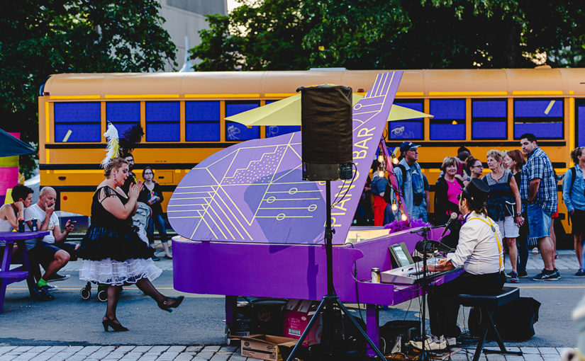 Acteur jouant sur un piano mauve avec motifs géométriques peints en jaune devant un kiosque aménagé dans un autobus scolaire