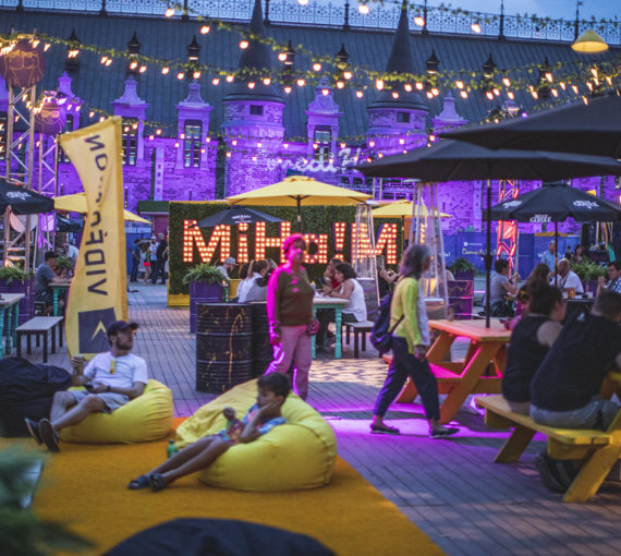 Visiteurs assis sous des parasols aux tables à pique-nique et dans des bean bags sur tapis de sol dans la zone Miha!m le soir