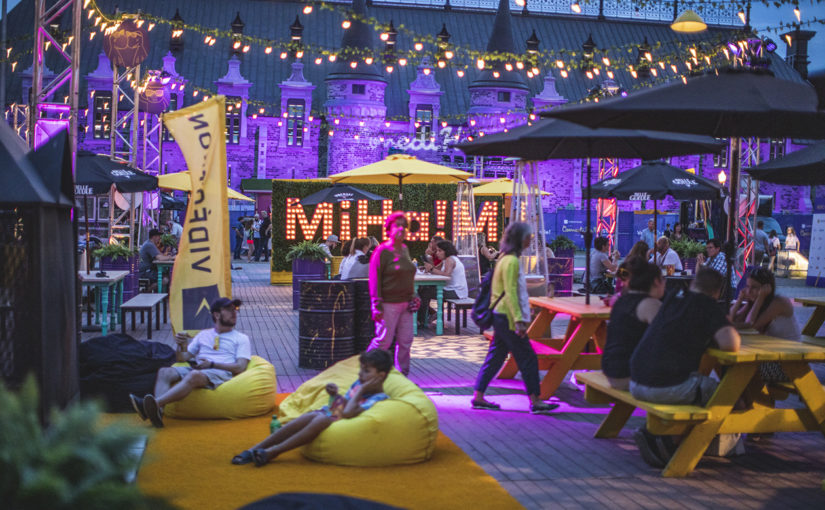 Visiteurs assis sous des parasols aux tables à pique-nique et dans des bean bags sur tapis de sol dans la zone Miha!m le soir