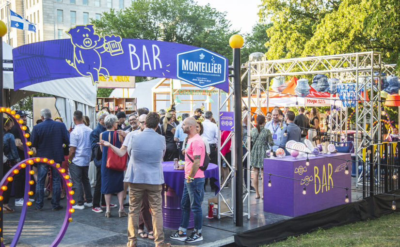 Rassemblement de gens debout autour de barils mauves recyclés profitant du festival dans l’espace bar sous un pont d’éclairage
