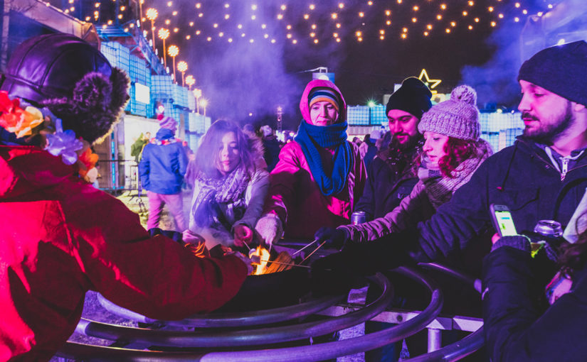 Festivaliers habillés de tuque, mitaines et foulard grillant des guimauves sur un brasero sous l’éclairage des string lights