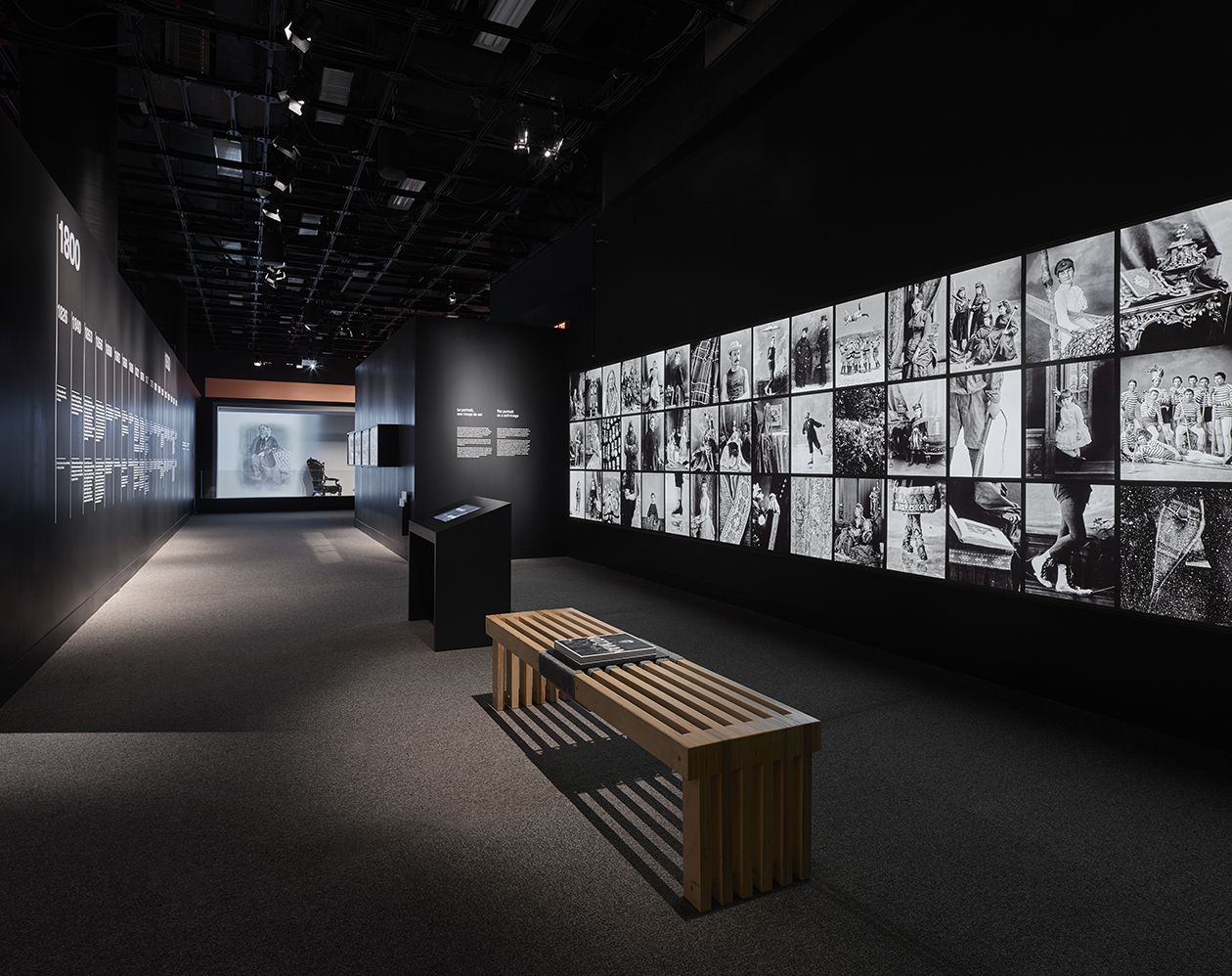 Zone d’exhibition avec un banc, une borne de consultation et une mosaïque de photos lumineuses noir et blanc sur une cimaise